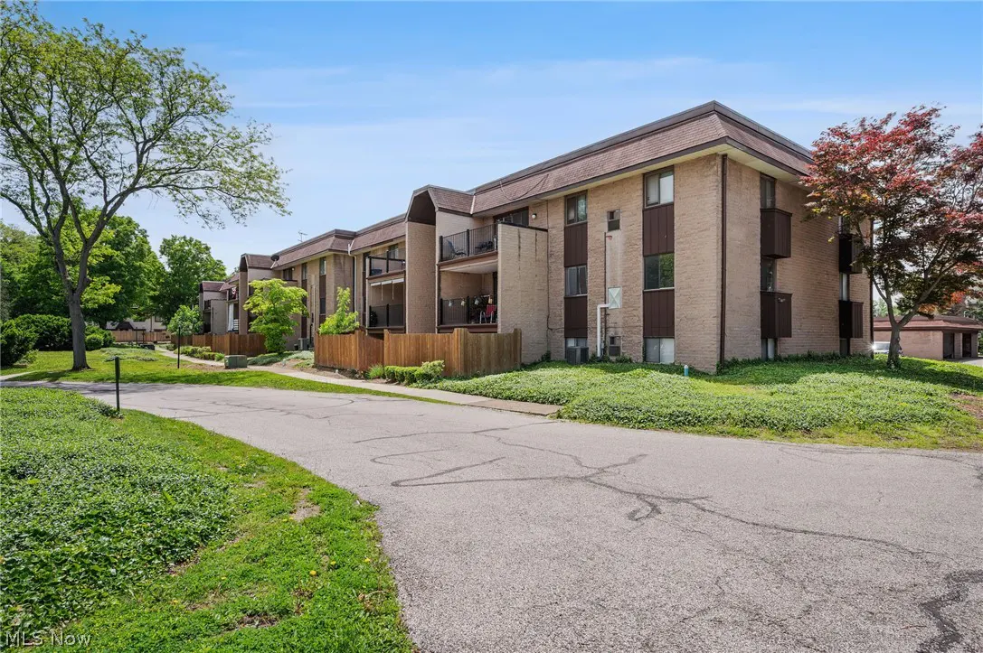 Exterior view of a tan brick apartment building with brown trim and balconies, surrounded by green grass and trees under a blue sky.