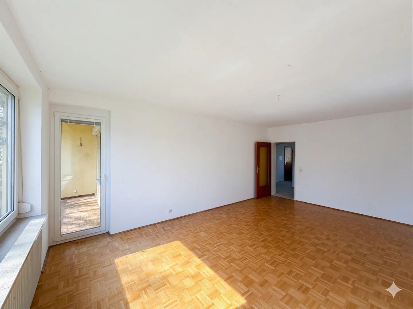 Bright, empty room with parquet floors, white walls, and a large window. A door leads to a balcony, and another to an adjacent room.