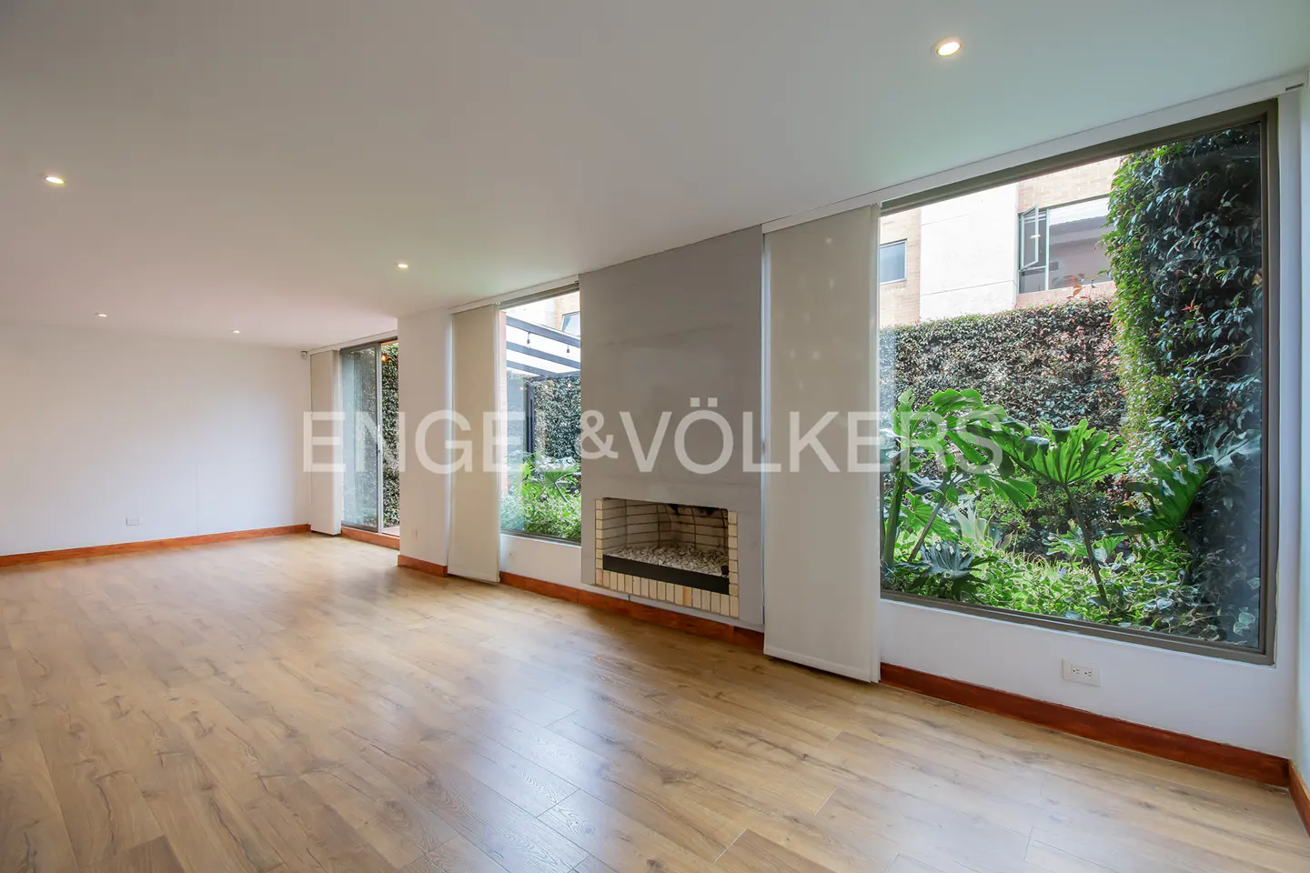 Bright, empty living room with wood floors, white walls, and a gray fireplace. Large windows overlook lush green gardens. Engel & Völkers logo visible.