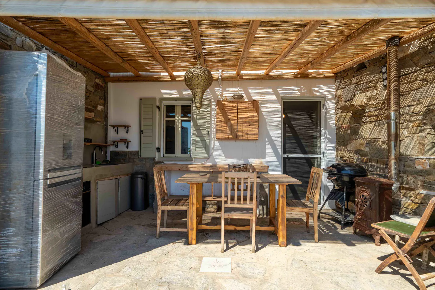 Outdoor dining area with wooden table and chairs under a thatched roof. A grill and wrapped refrigerator are also visible.