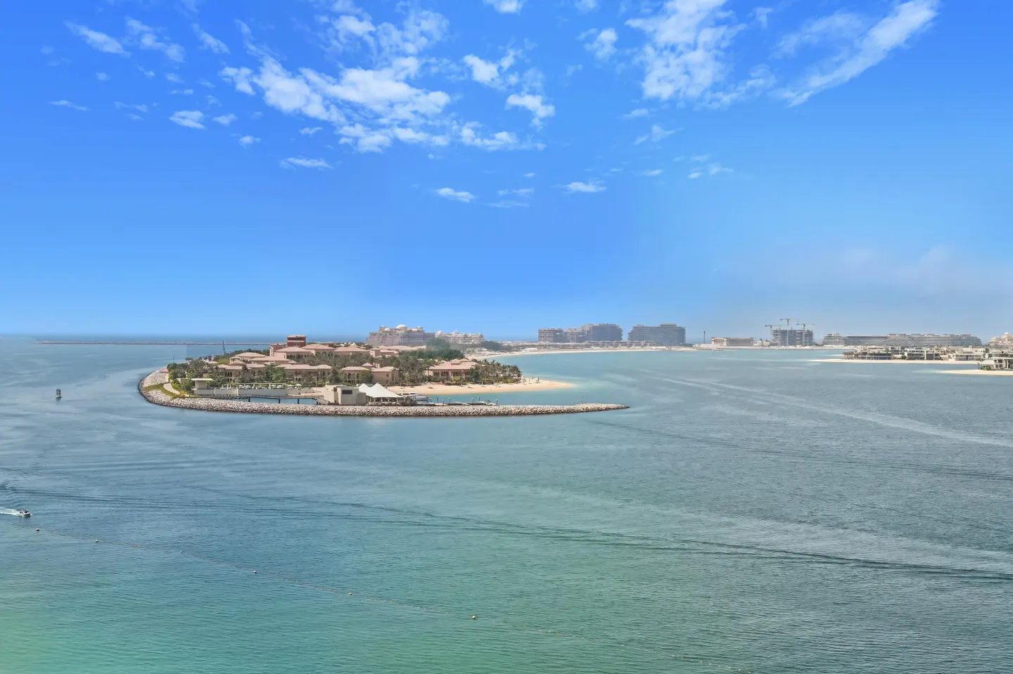 Island view of Dubai's Palm Jumeirah with luxury villas, sandy beach, and turquoise water under a blue sky with scattered clouds.