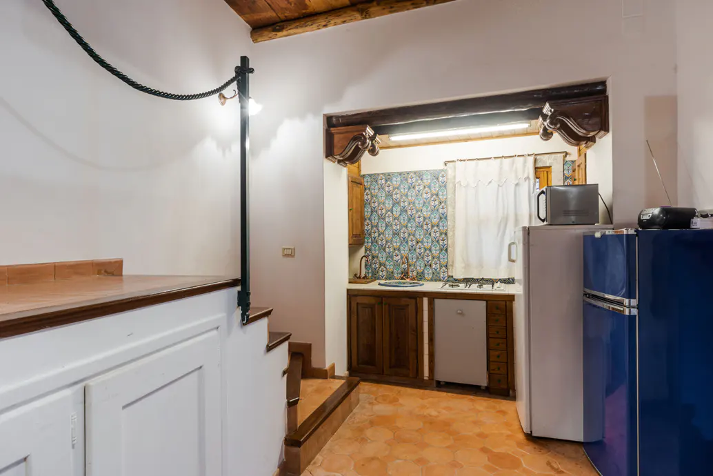 A cozy kitchen with terracotta floors, wooden cabinets, and a blue vintage refrigerator. Stairs lead up to a counter with white cabinets.