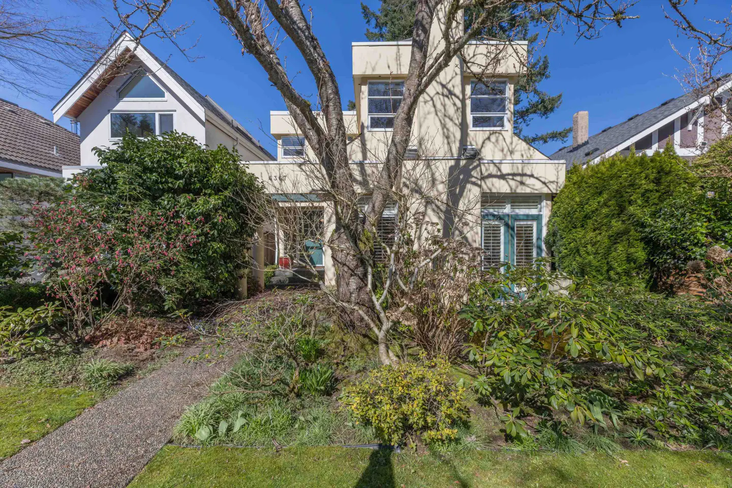 A two-story beige house with a tree in front, surrounded by green bushes and a stone path leading to the entrance.