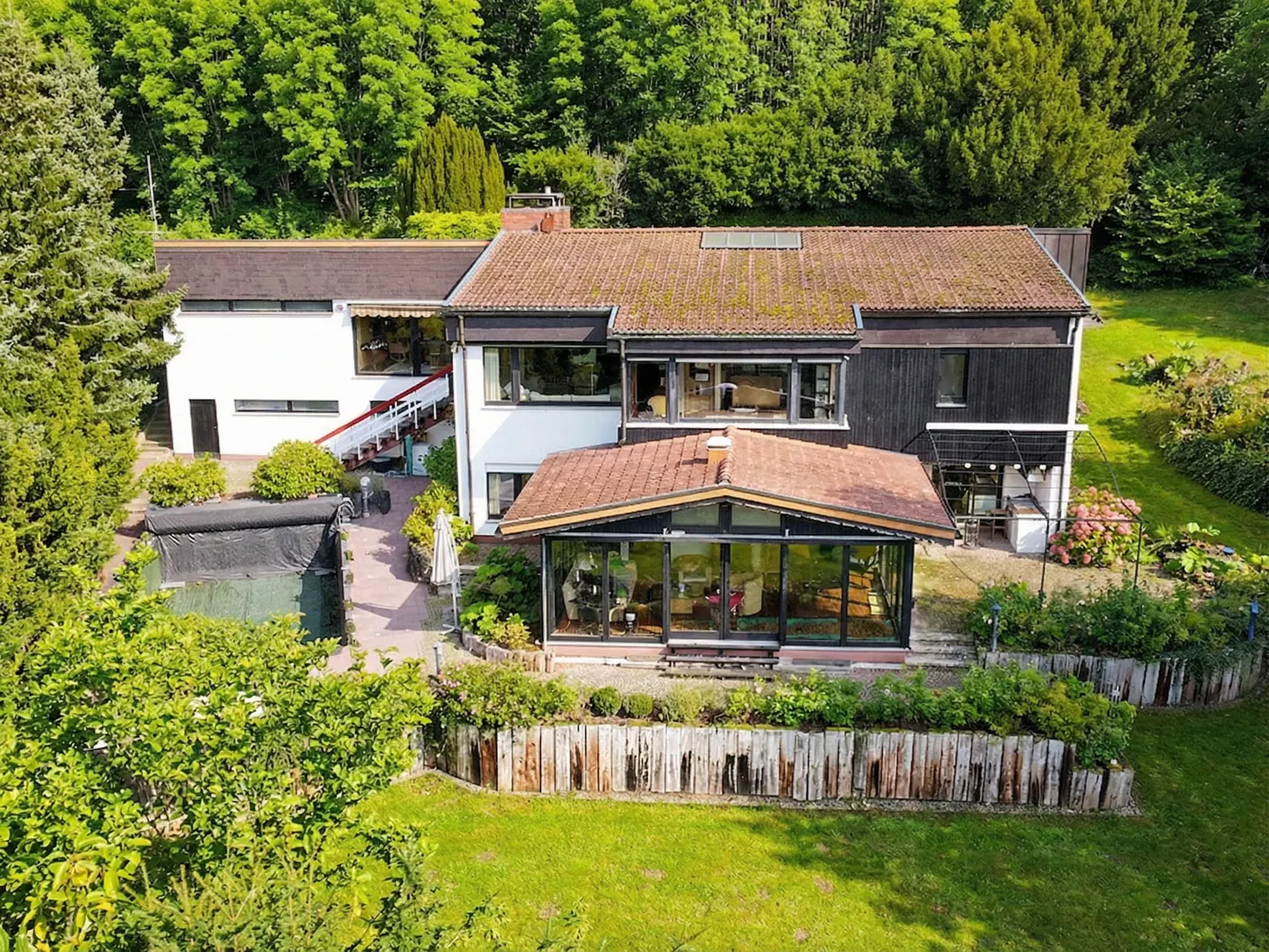 Aerial view of a modern house with a brown roof, white and black walls, a glass conservatory, and a green lawn surrounded by trees.