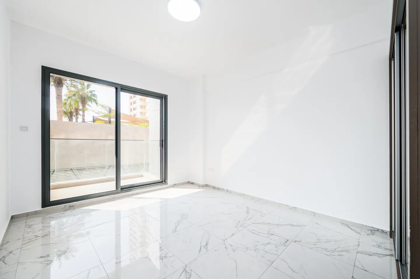 Bright, empty room with white walls and marble tile floor. Sliding glass doors lead to a balcony with palm tree views.