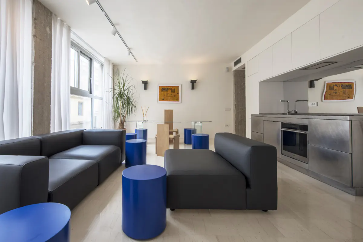 Bright, modern apartment with black sofas, blue cylindrical stools, and a glass-topped dining table. Stainless steel kitchen in the background.