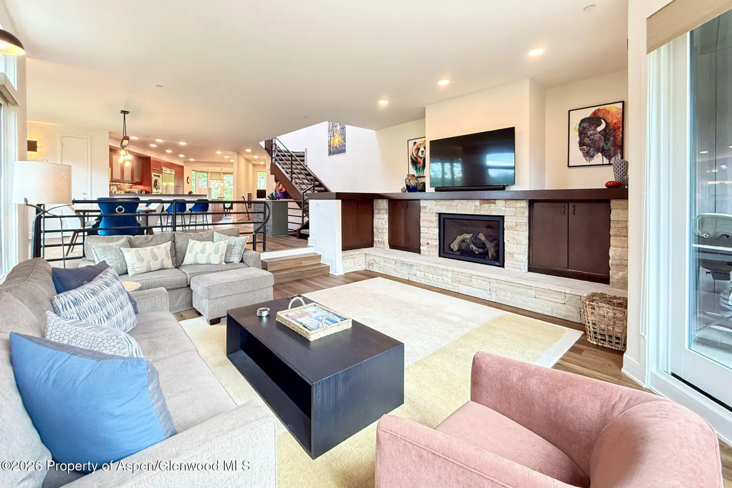 Modern living room with gray sectional, pink armchair, stone fireplace, TV, and open view to kitchen and staircase.