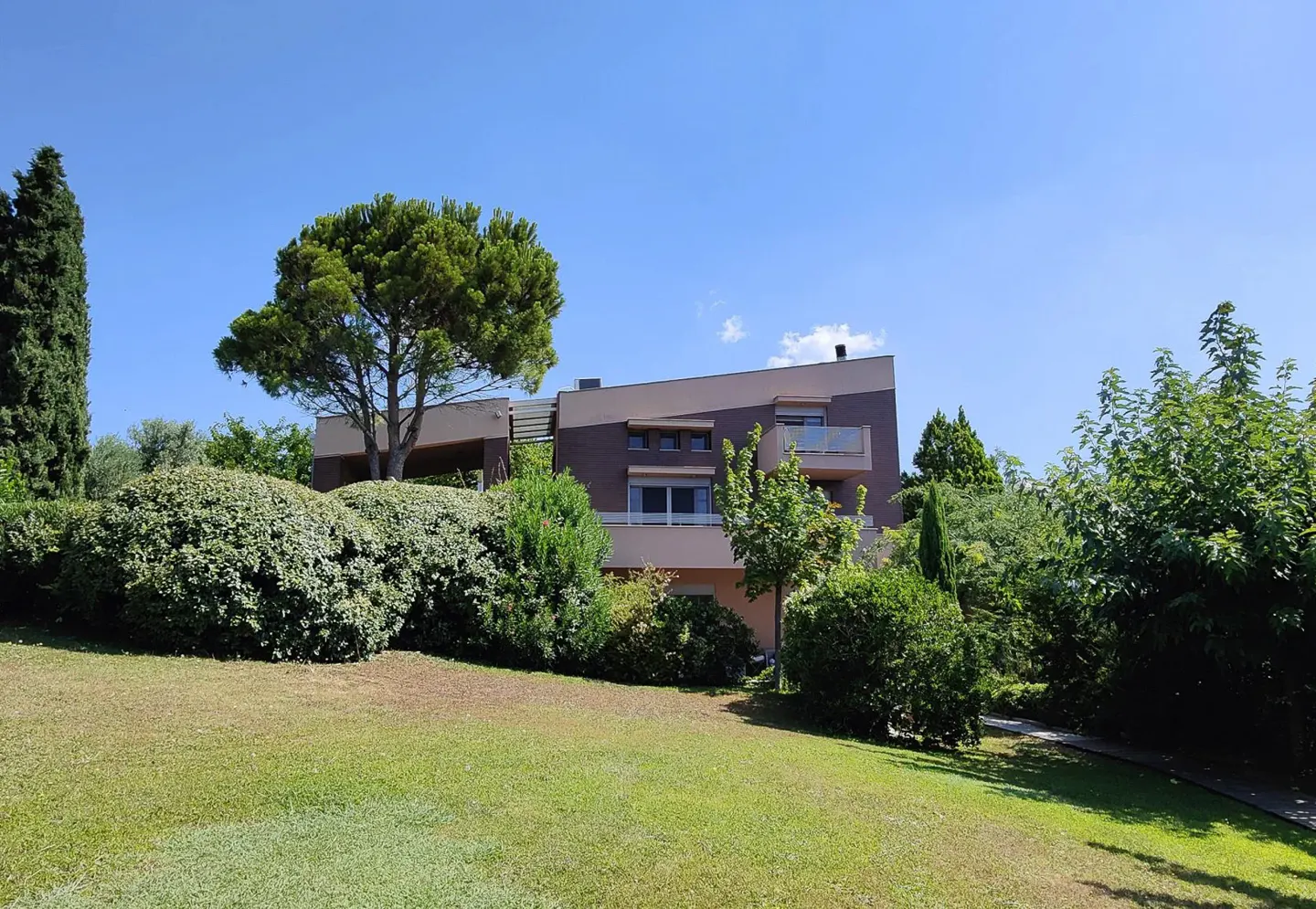 Modern two-story house with brown and beige exterior, surrounded by lush green trees and a manicured lawn under a blue sky.
