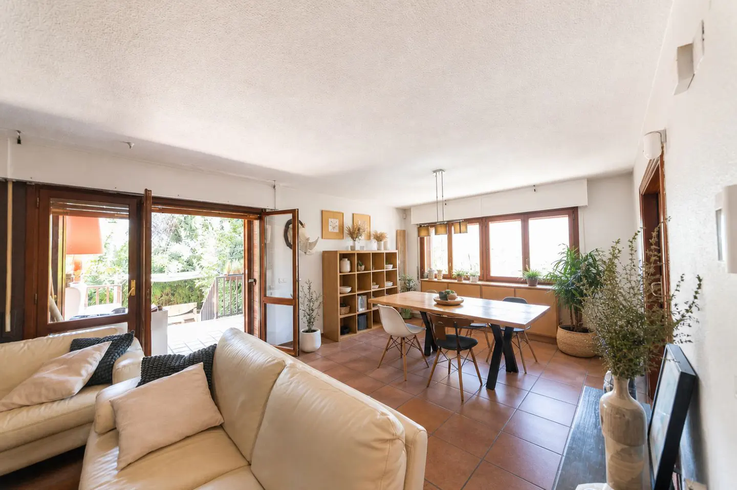 Bright living room with a cream sofa, dining table with chairs, and open doors to a balcony with greenery.