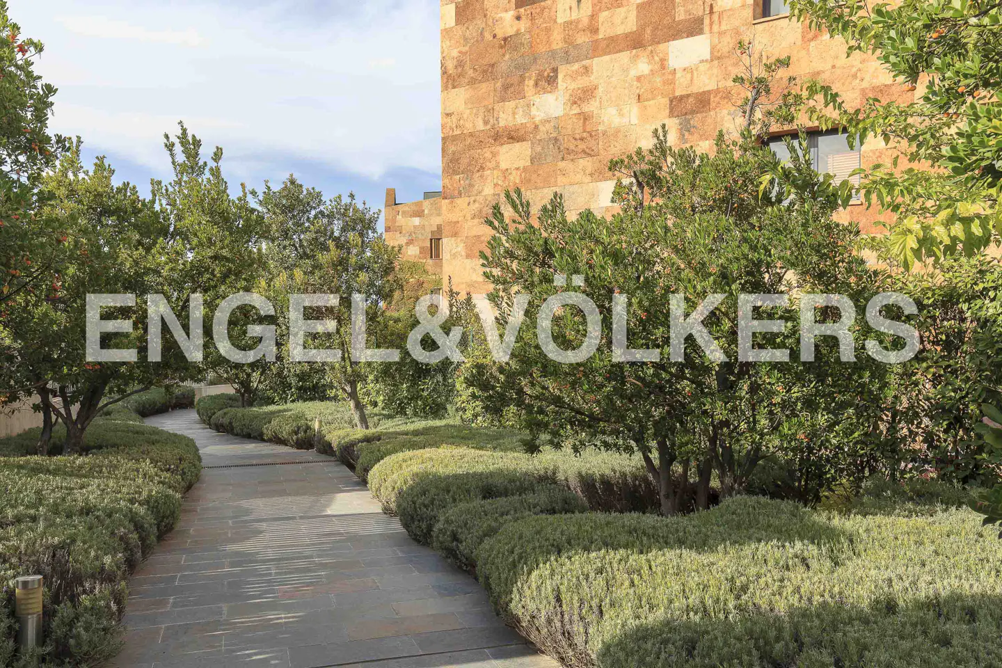 A stone path winds through lush green landscaping toward a large, tan stone building under a blue sky.