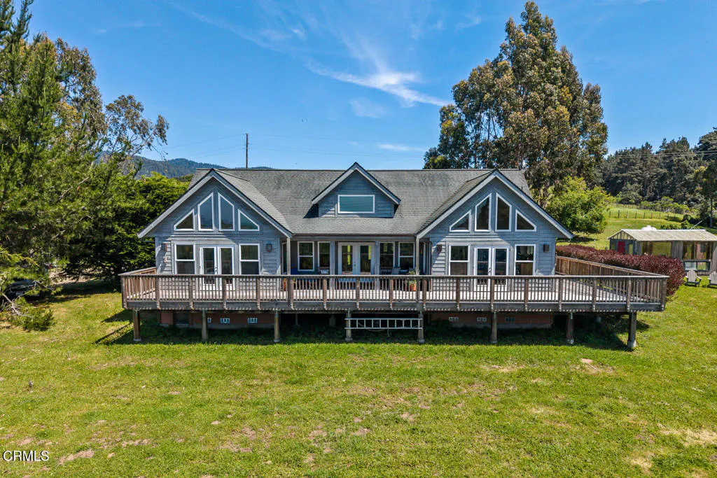 A gray house with a large wooden deck sits on a green lawn under a blue sky.