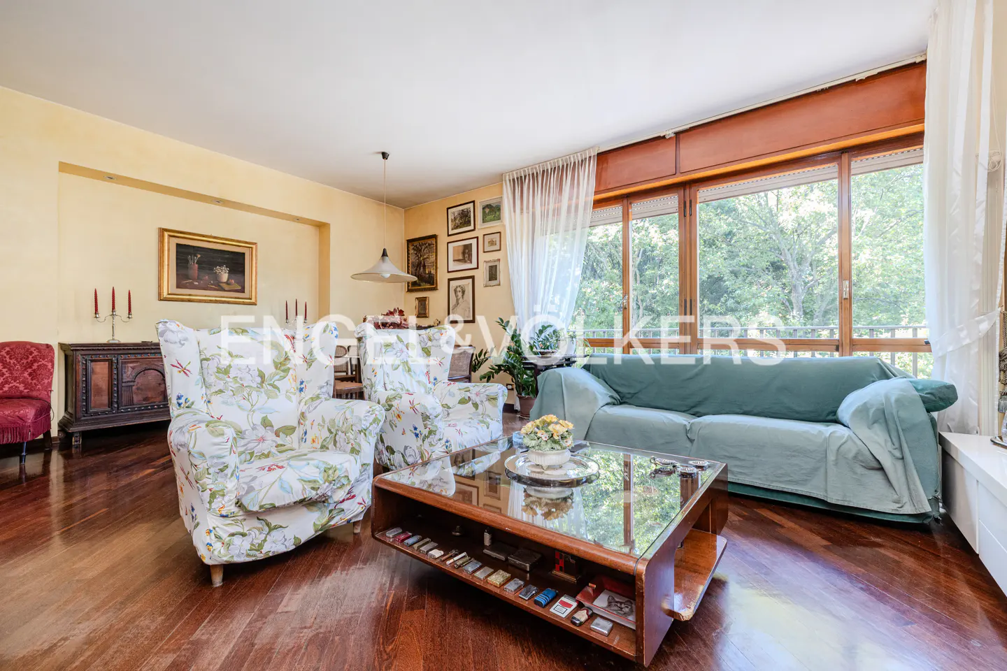 Living room with two floral armchairs, a green sofa, a glass coffee table, and large windows overlooking trees.