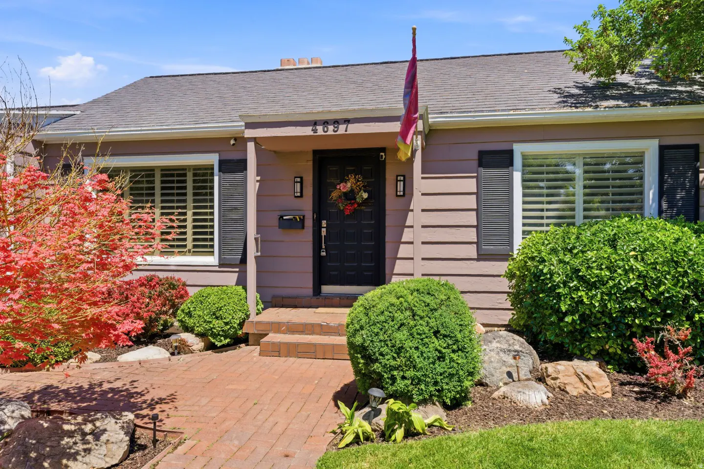 Exterior of a single-story house with brown siding, black shutters, and a brick walkway. A flag hangs above the black front door.