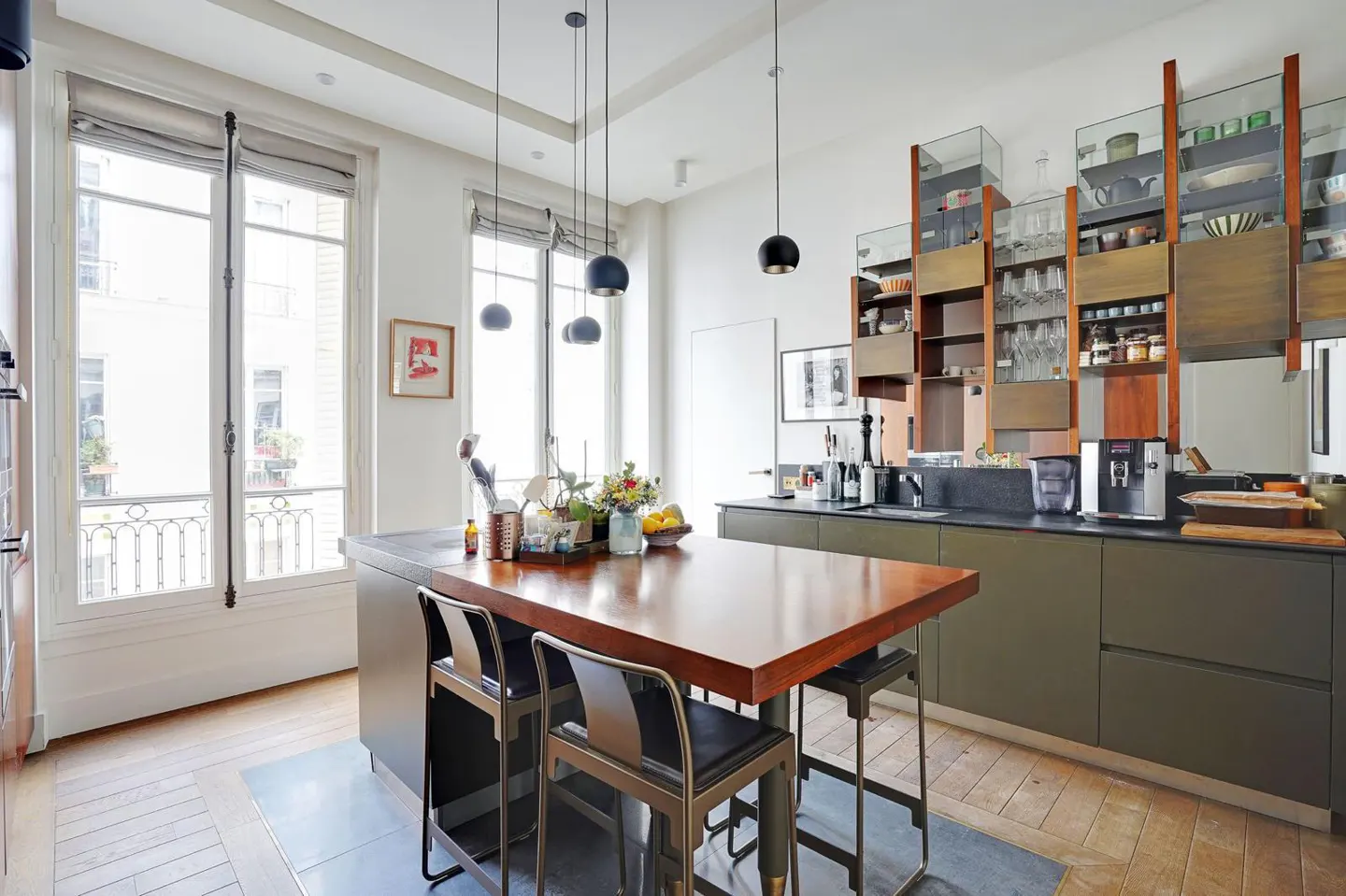 A modern kitchen with a wood island, black stools, and olive green cabinets. Tall, glass-fronted cabinets display dishware. Large windows let in natural light.