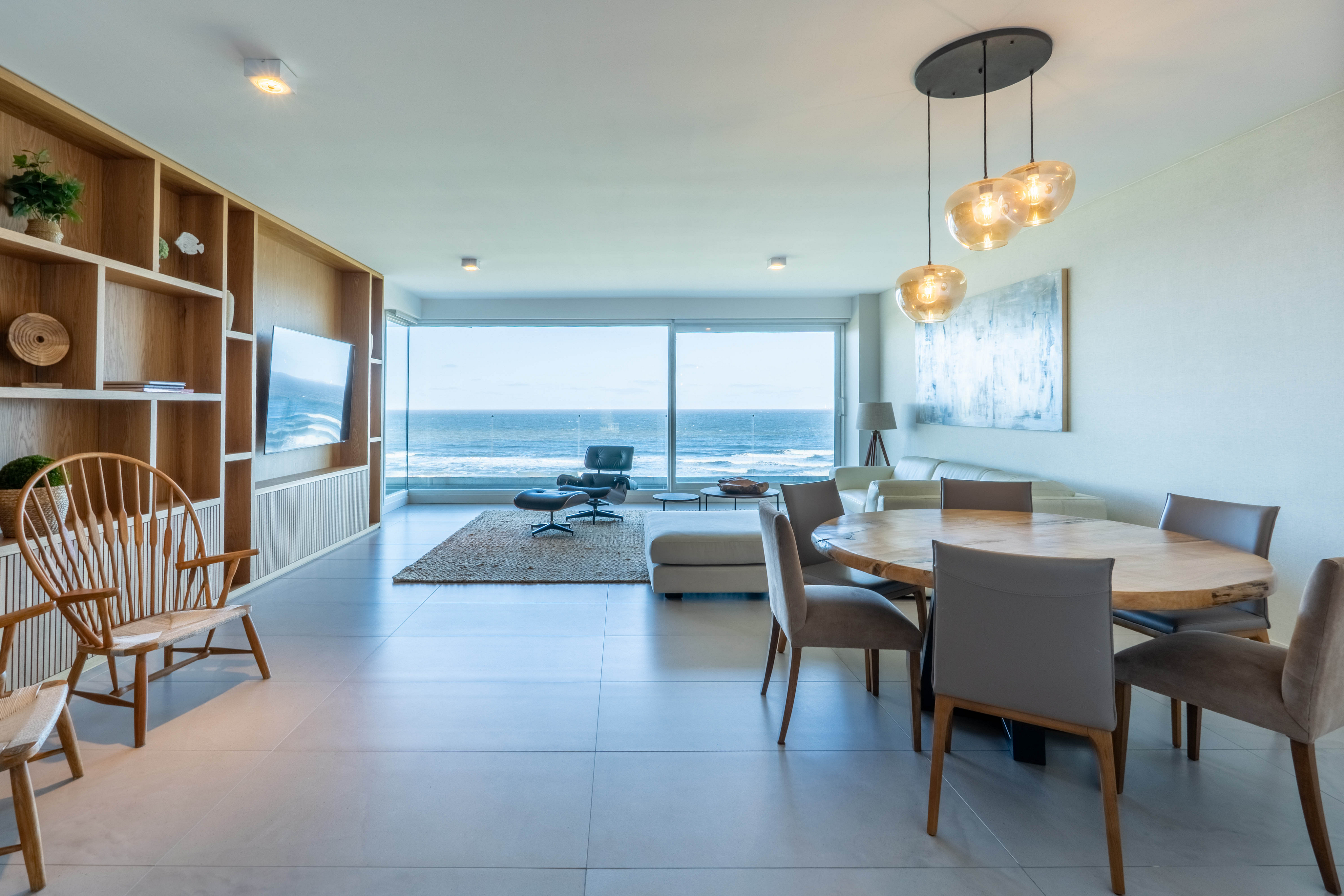 Modern open-plan living room with dining area, wooden shelving, and floor-to-ceiling windows overlooking the ocean.