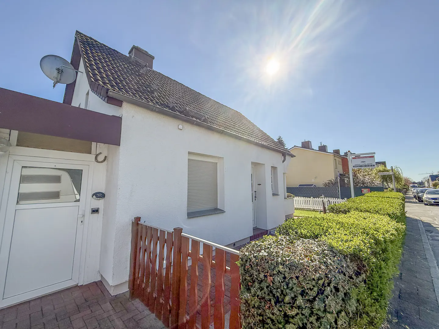 Exterior view of a one-story white house with a brown roof, a satellite dish, and a small wooden fence.