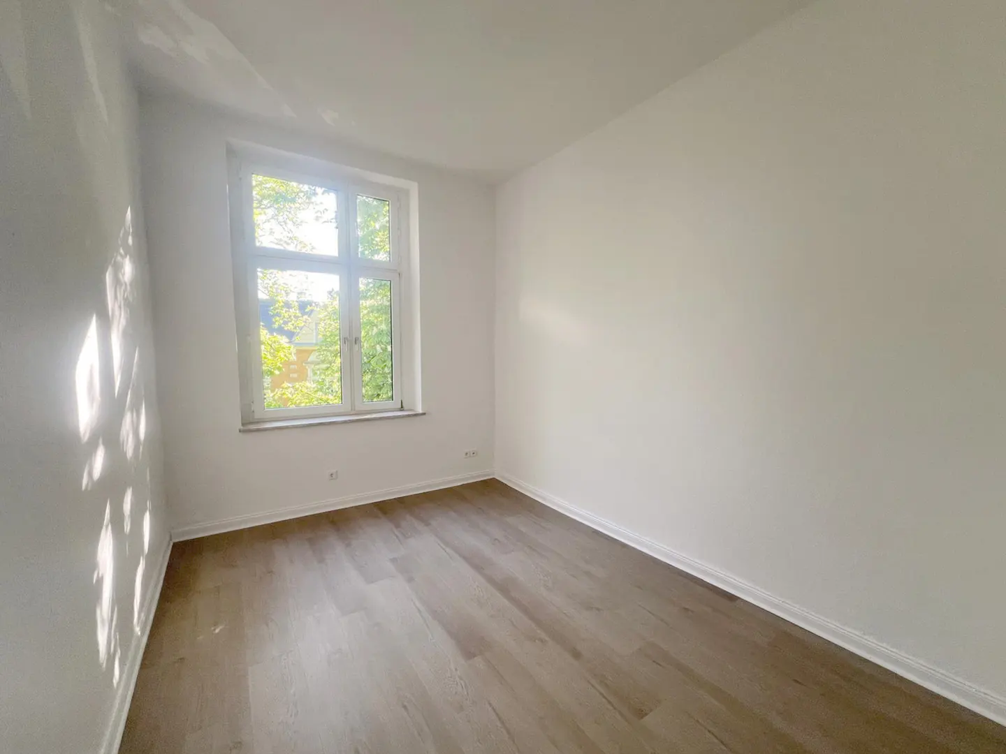 Bright, empty room with light wood floors and white walls. A window lets in natural light, showing green trees outside.