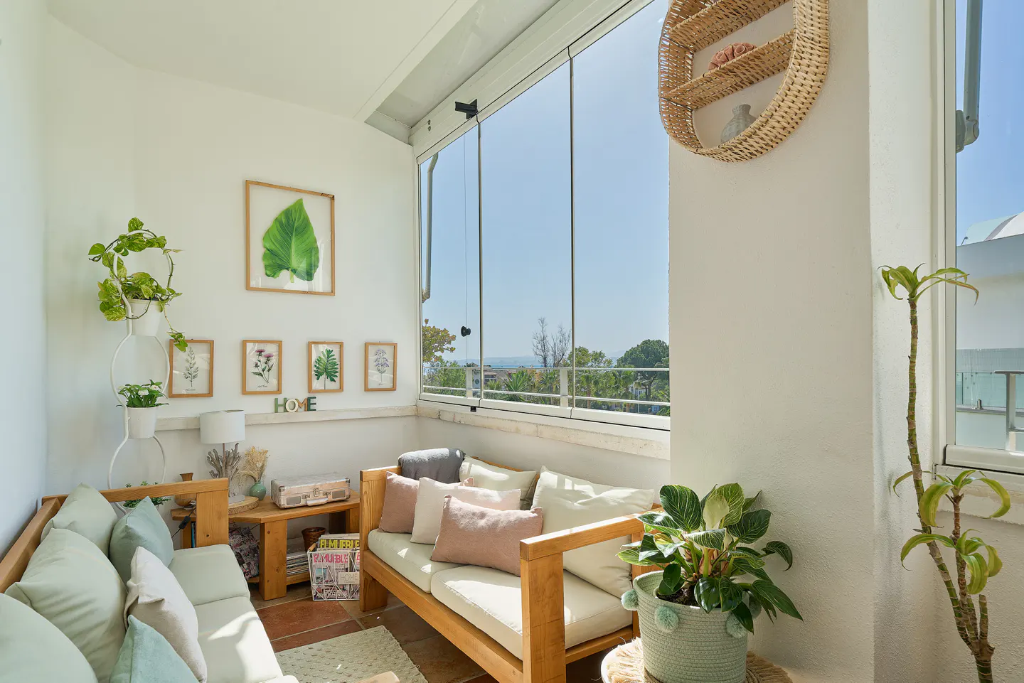 Bright sunroom with wooden sofas, pastel pillows, and potted plants. Framed botanical prints hang on the white wall. Glass doors offer a view of trees and sky.