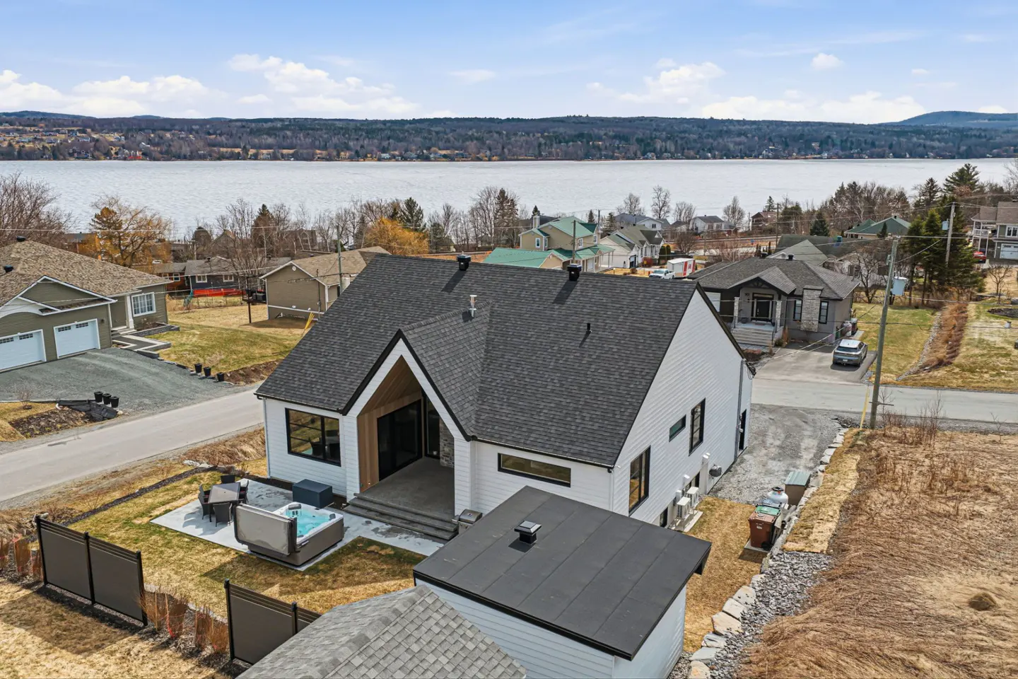 Aerial view of a modern white house with a black roof, a hot tub, and a lake in the background.