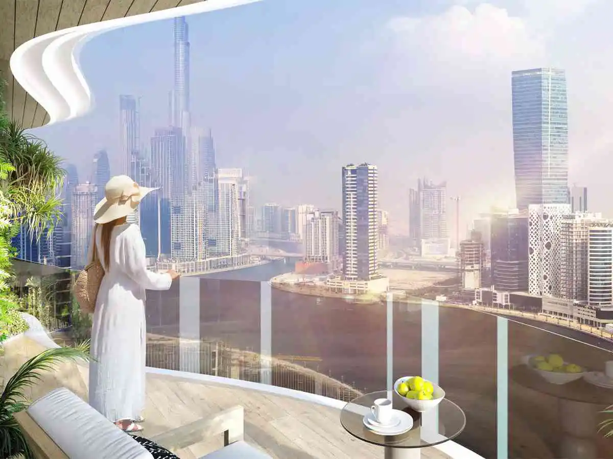 A woman in white stands on a balcony overlooking the Dubai skyline. A table with fruit and a cup sits nearby.