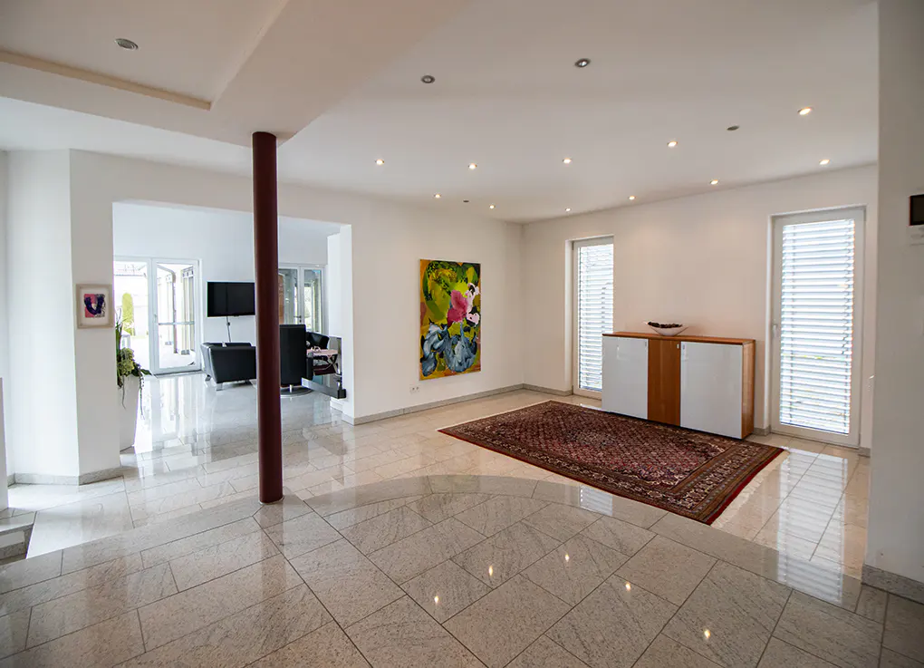 Bright, open-concept living space with marble floors, white walls, and recessed lighting. A red rug sits near a cabinet and windows. An abstract painting hangs on the wall.
