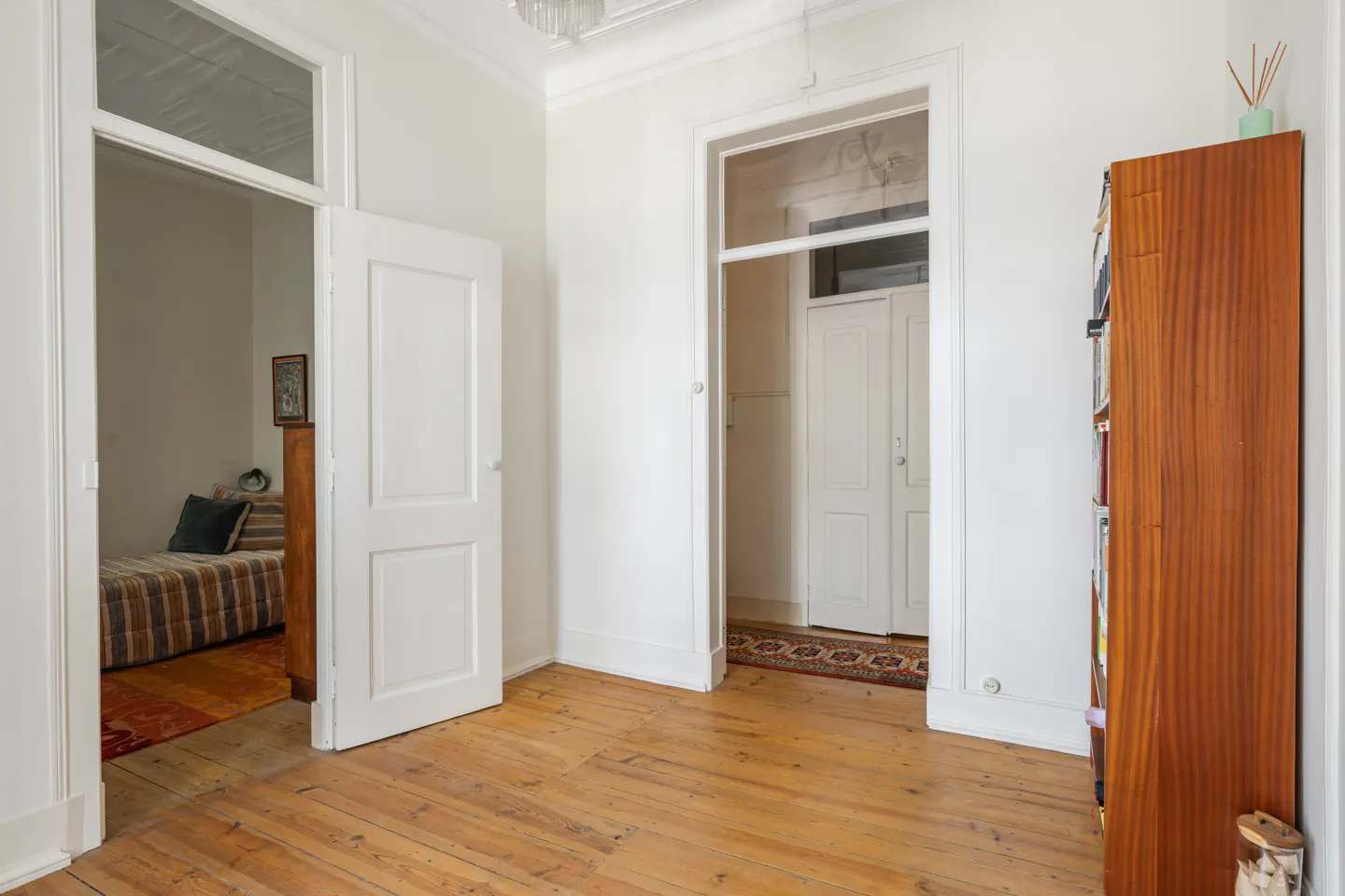 Bright room with wood floors, white walls, and two doorways. One leads to a bedroom, the other to a closet. A tall, narrow bookcase stands against the right wall.