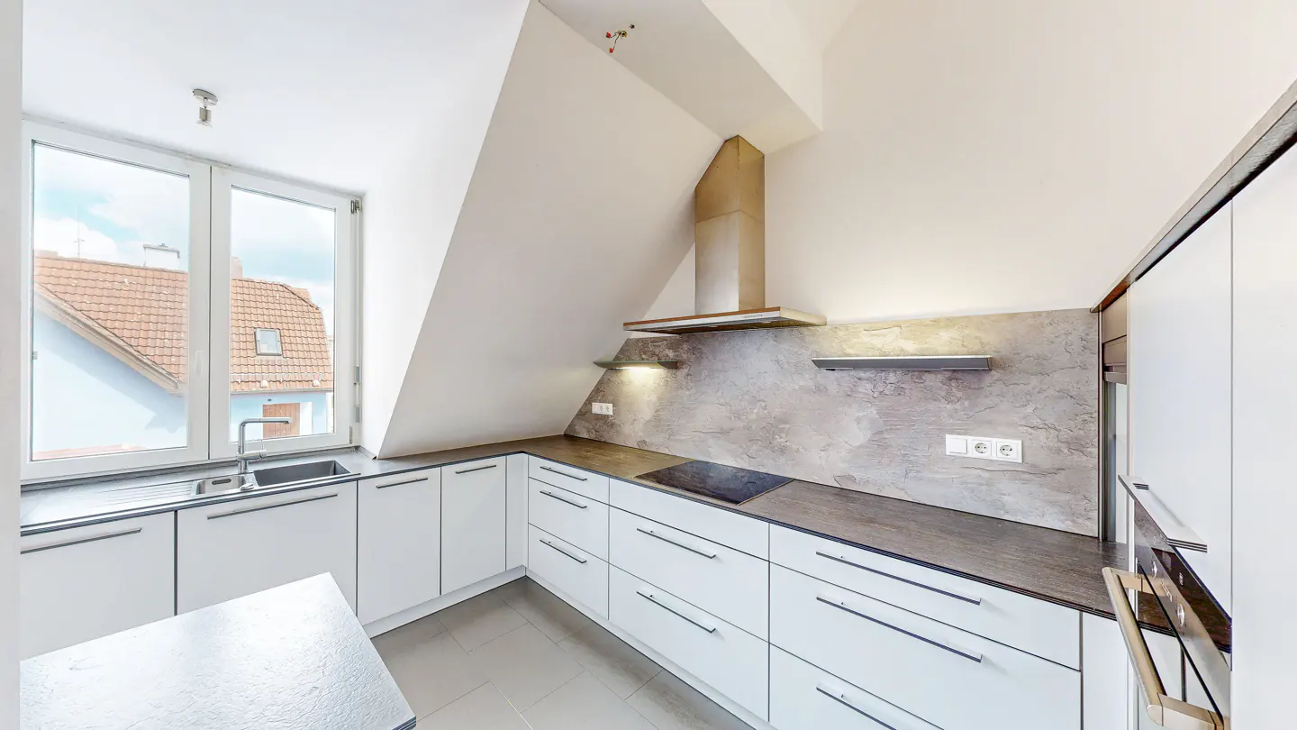 Modern attic kitchen with white cabinets, dark countertops, textured backsplash, and a window overlooking tiled rooftops.