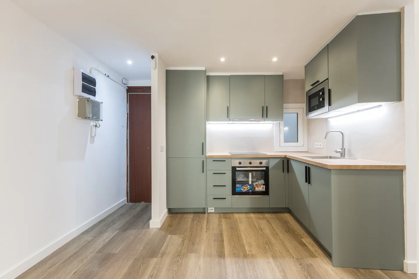 A modern kitchen with light green cabinets, wood countertops, and wood-look flooring. A stainless steel microwave and oven are visible.