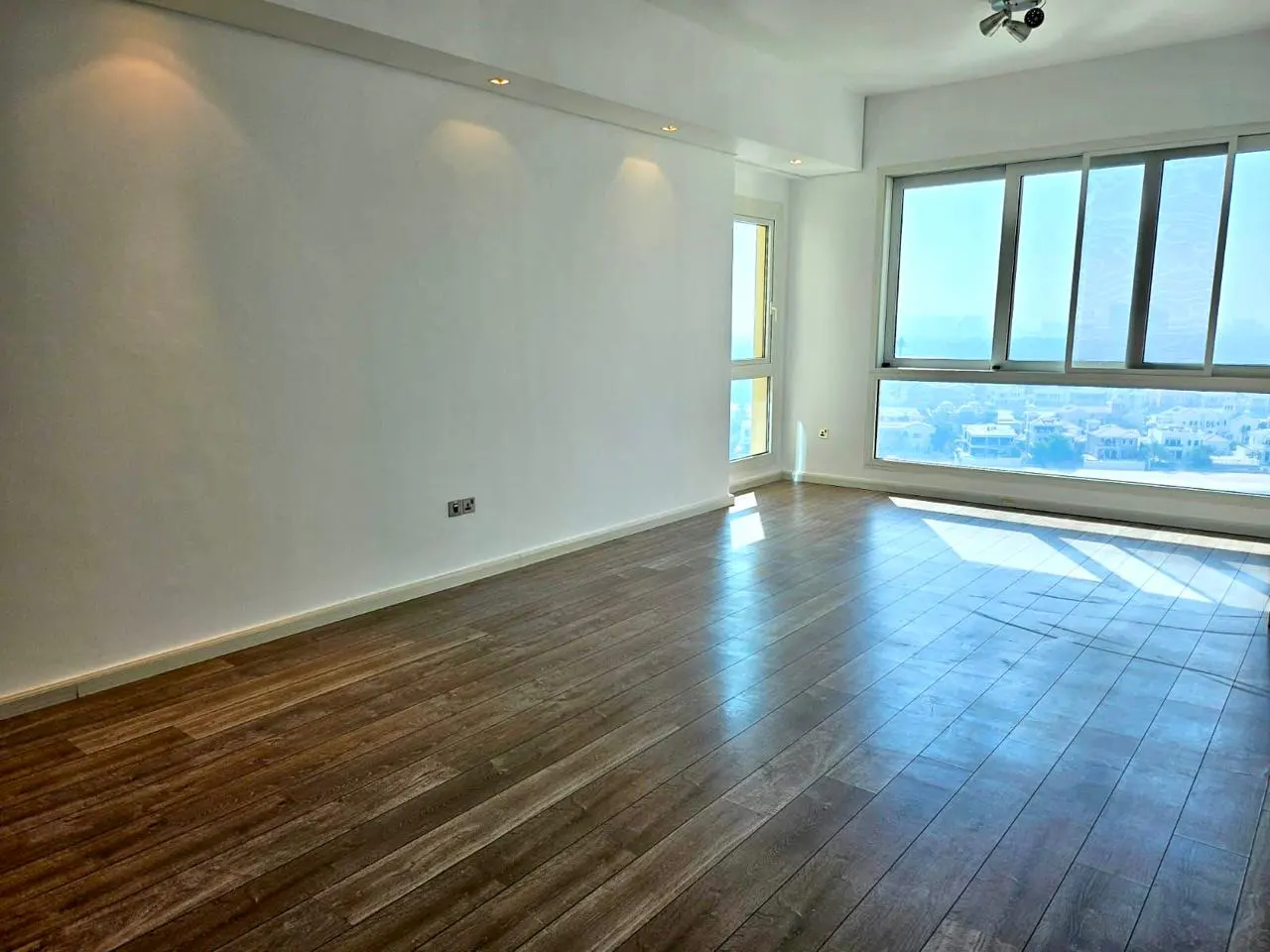 Bright, empty room with wood floors, white walls, and large windows showing a city and ocean view.