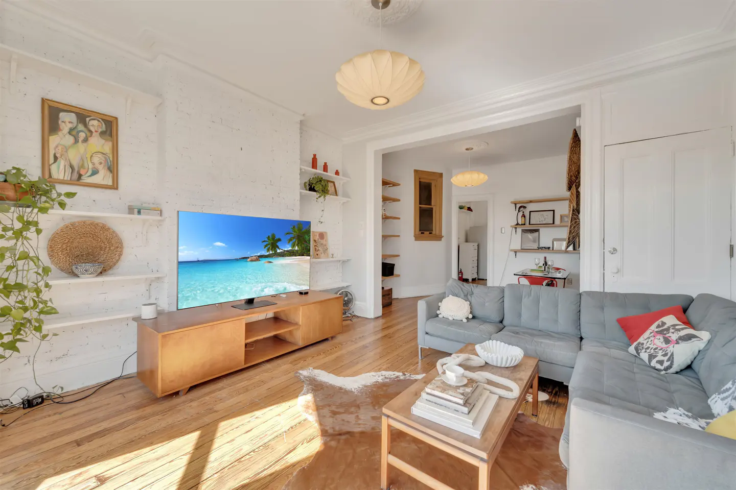 Bright living room with hardwood floors, a gray sectional sofa, and a wooden TV stand displaying a beach scene. White walls and shelves add to the airy feel.