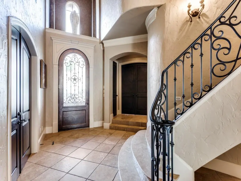 Elegant foyer with tile floors, dark wood doors, and a wrought iron staircase. A statue is visible above the front door.