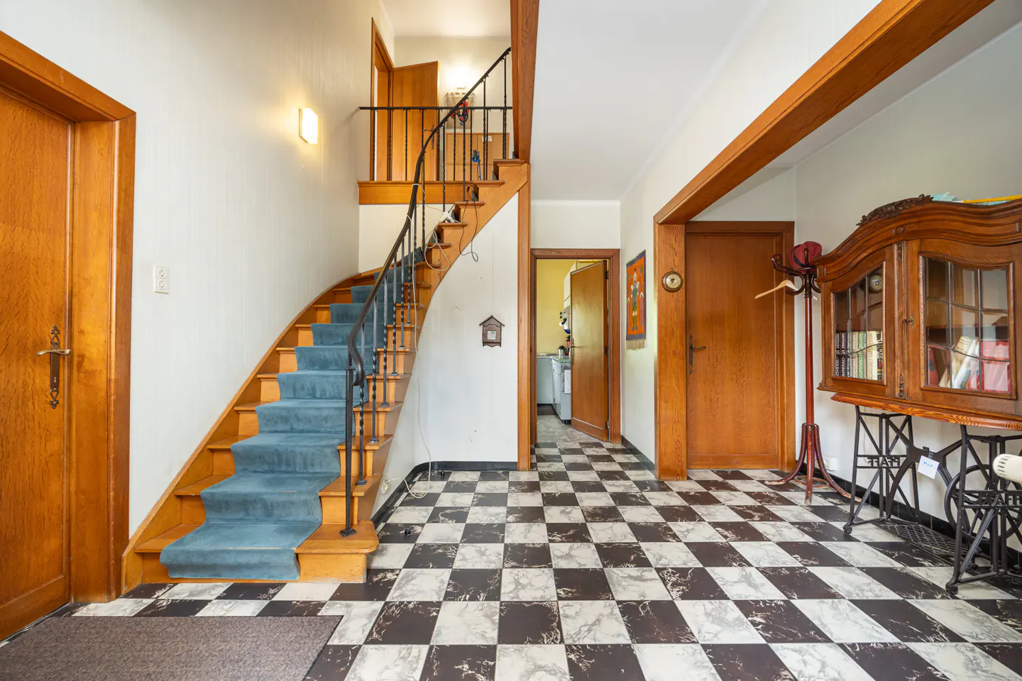 Entryway with black and white checkered floor, blue carpeted staircase, wooden doors, and a vintage sewing machine cabinet.