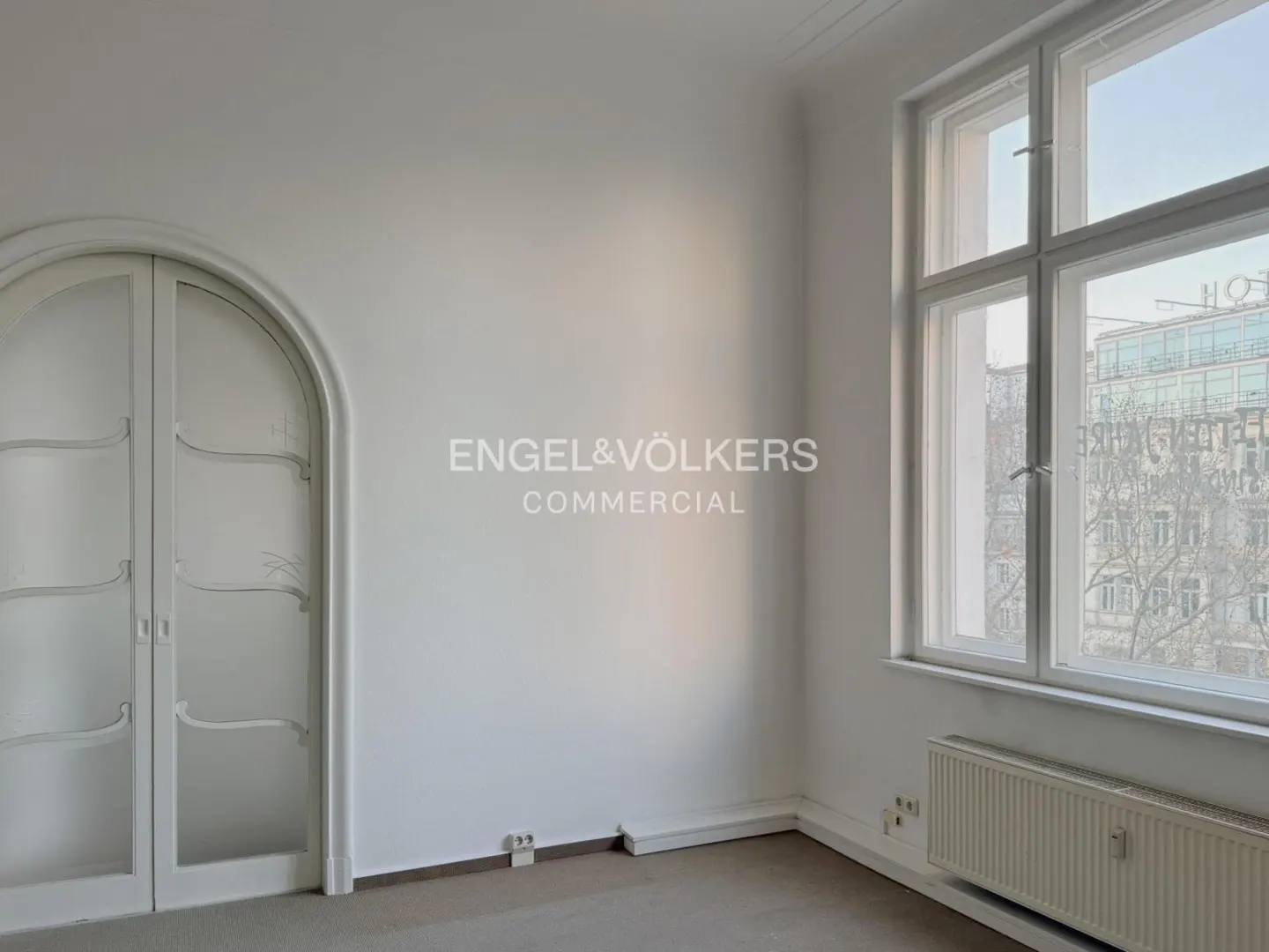 Bright, empty room with white walls, arched glass doors, and a large window showing a city view. A radiator sits beneath the window.