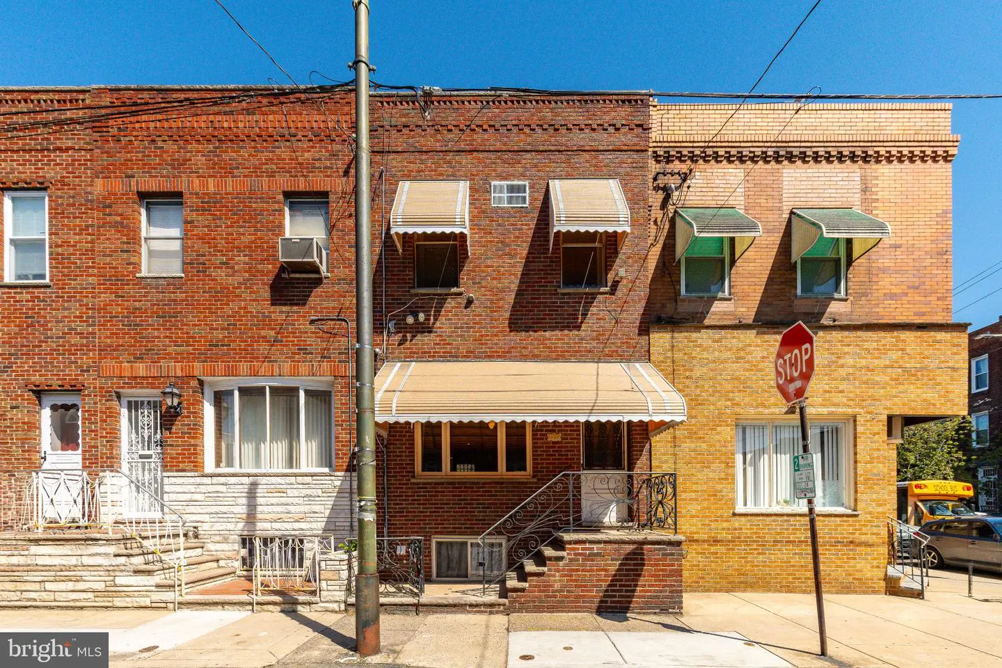 Row houses with brick facades and awnings line a city street under a clear blue sky. A stop sign is visible.