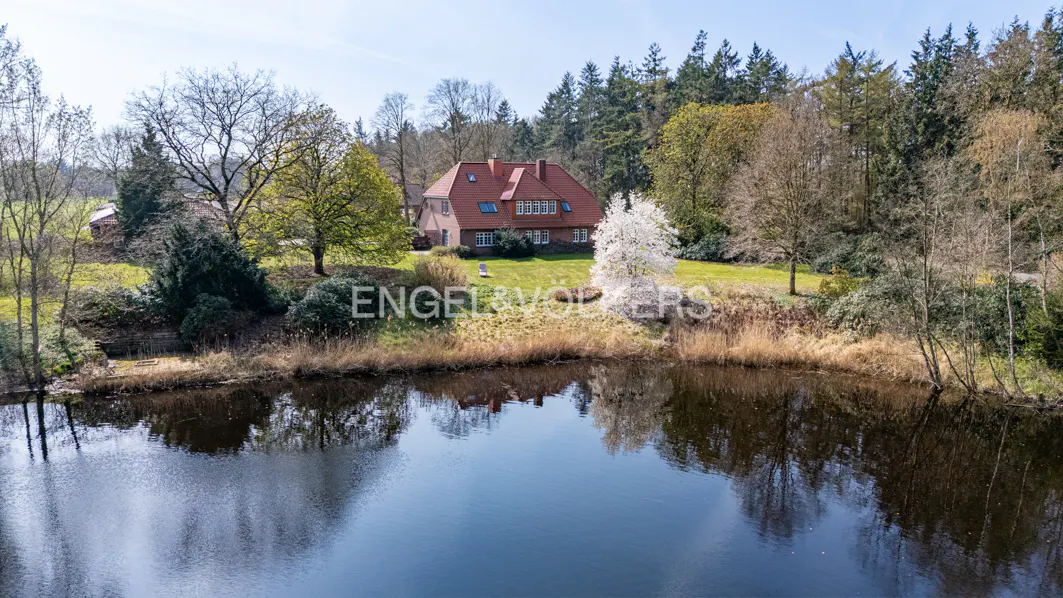 Aerial view of a brick house with a red tile roof, surrounded by trees, reflected in a pond.