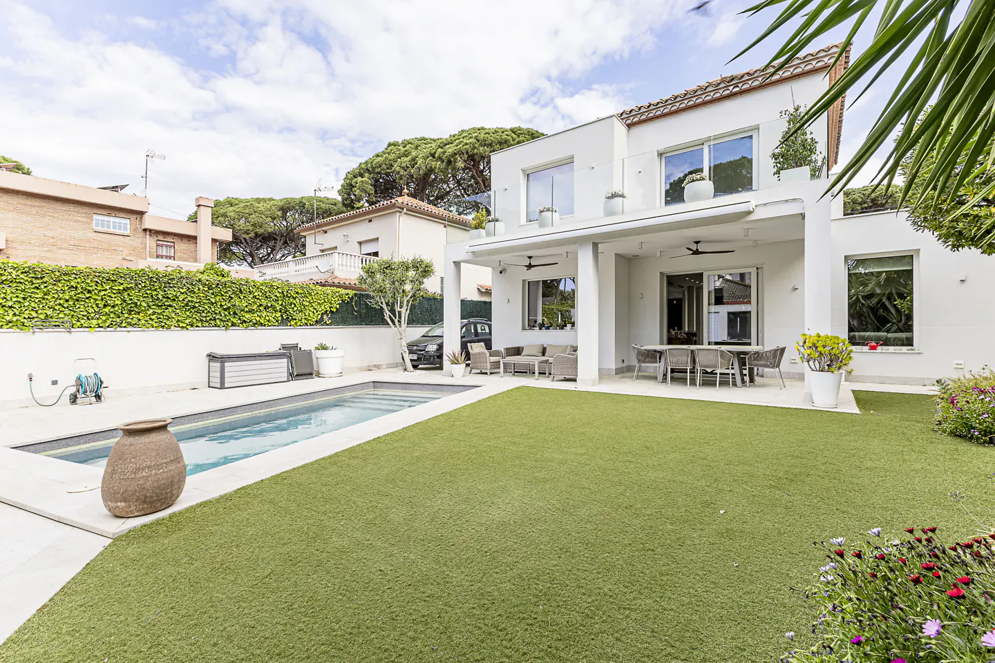 Backyard view of a modern white house with a pool, green lawn, patio furniture, and flower beds.