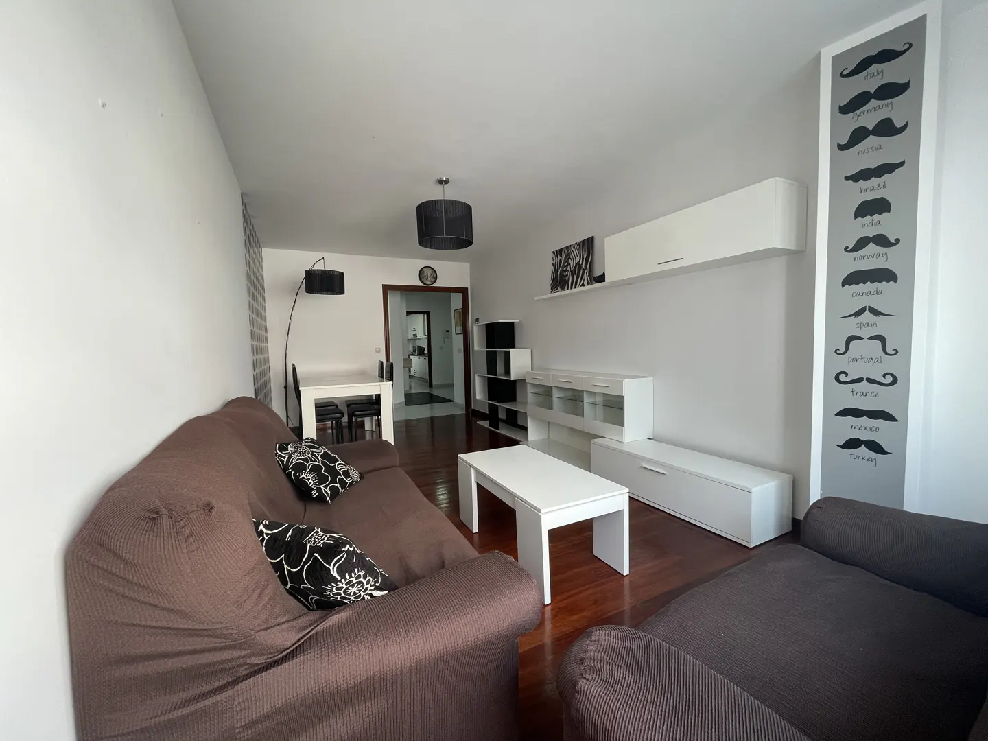 Living room with brown sofa, white furniture, and a wall decoration with mustache styles from different countries.