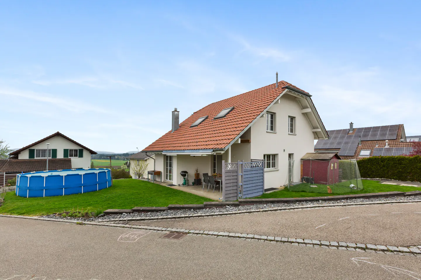 A white two-story house with a red tile roof, a blue above-ground pool, and green grass under a blue sky.