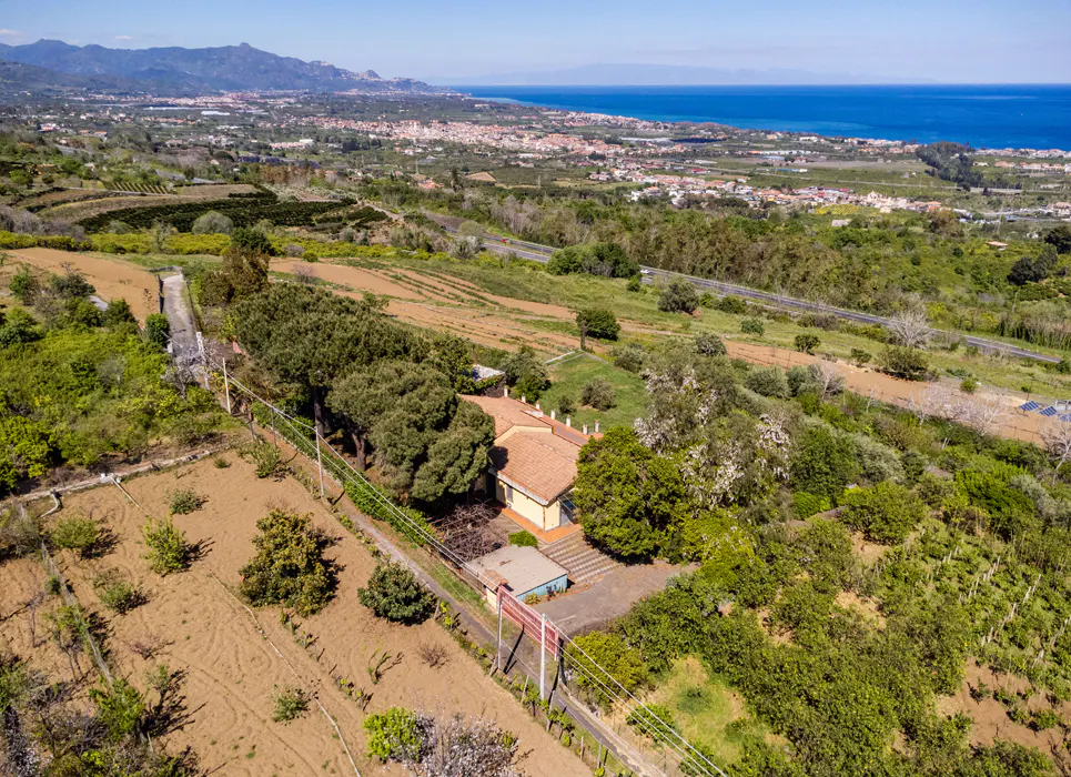 Aerial view of a yellow house with a red tile roof, surrounded by green trees and fields, with a coastal town and blue sea in the background.
