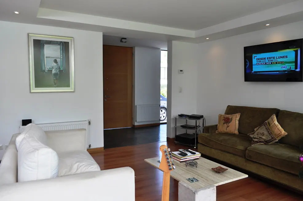 Living room with white walls, wood floors, and recessed lighting. A brown couch and white sofa face a TV. A painting hangs on the wall.