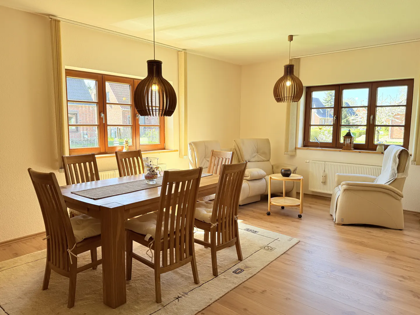 A bright living room with a wooden dining table, chairs, and two pendant lights. Beige sofas and a chair are in the background. Two windows let in natural light.