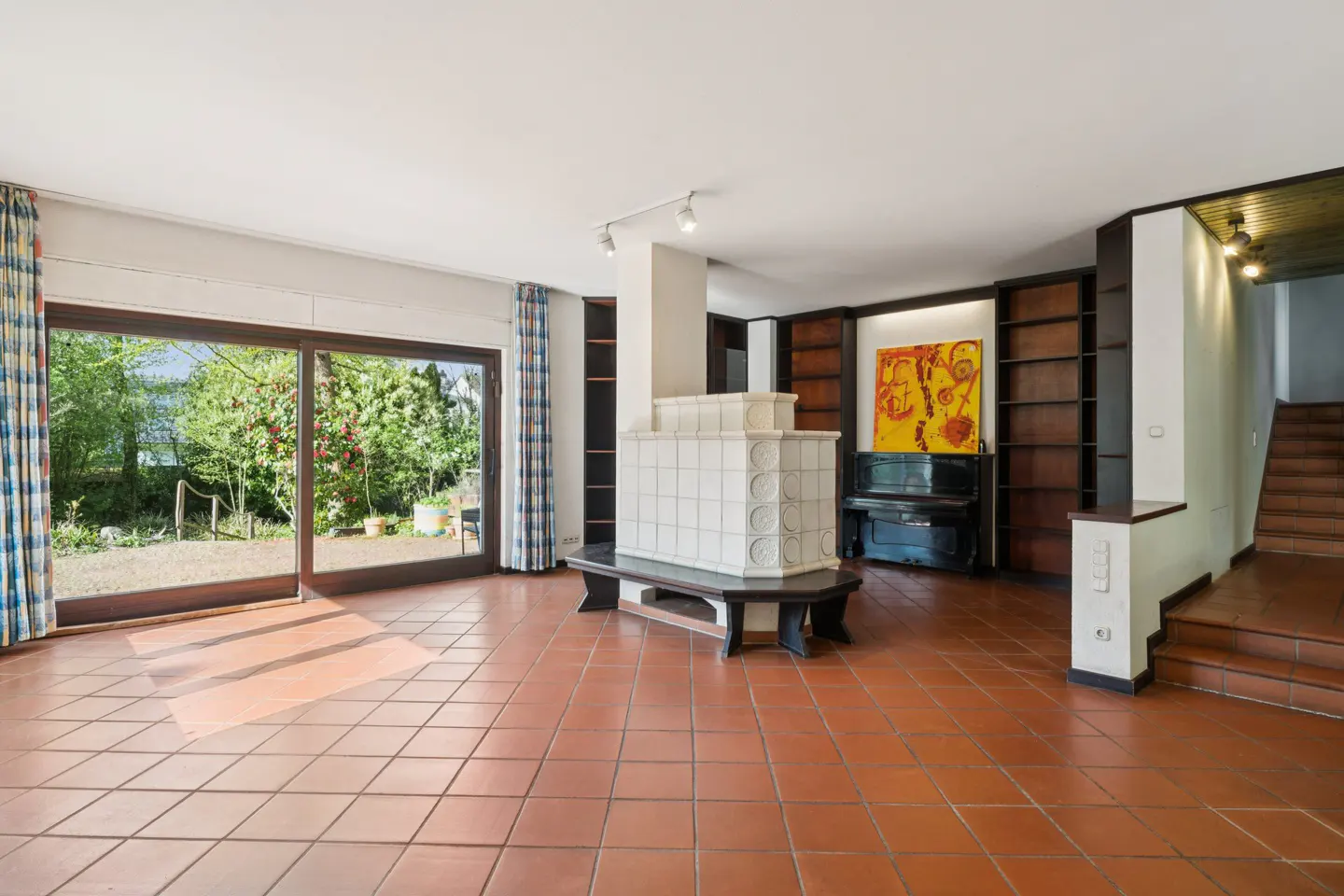Bright living room with terracotta tile floor, white tiled fireplace, piano, and sliding glass doors to a green garden.