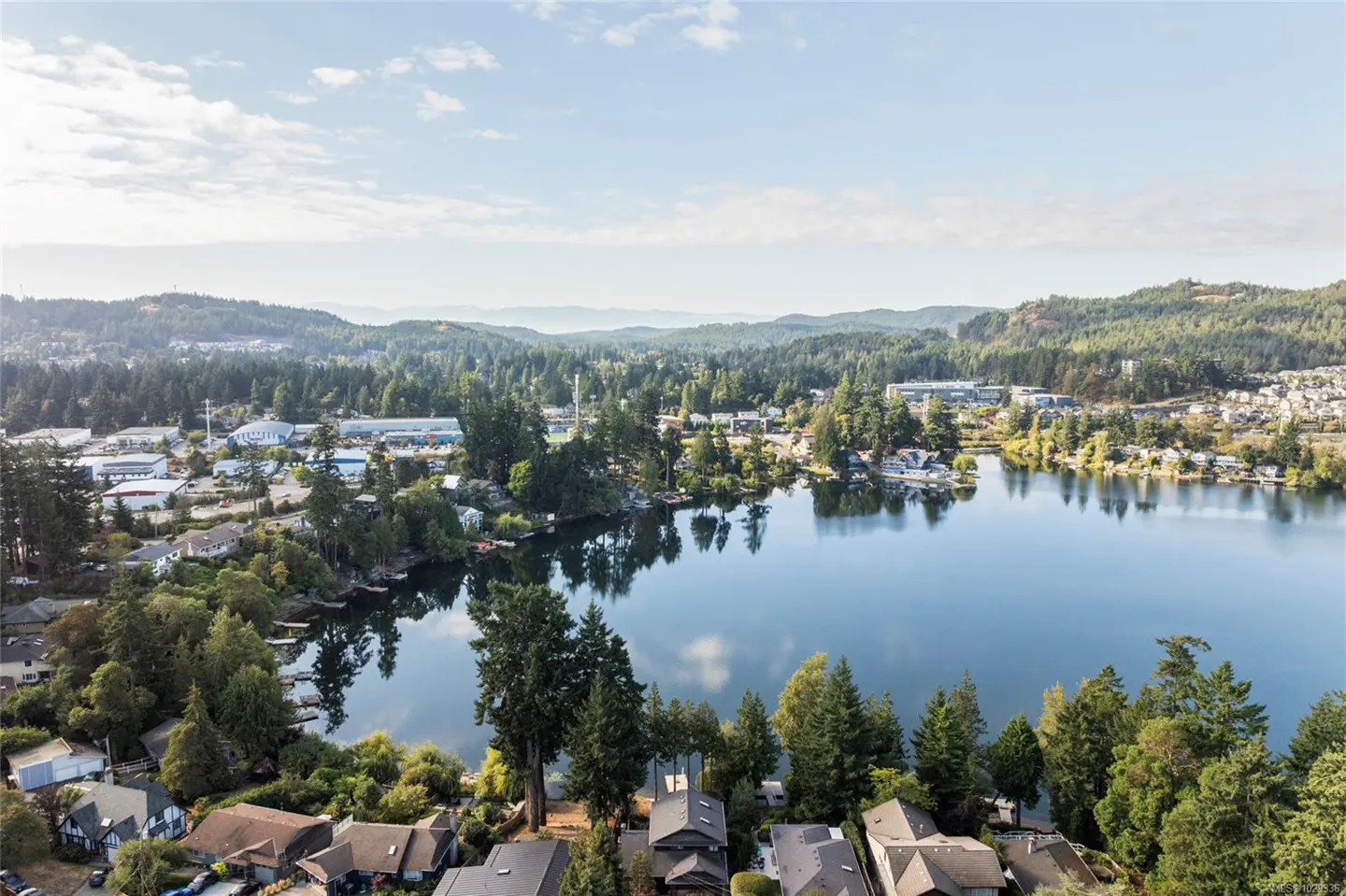 Aerial view of a blue lake surrounded by green trees, houses, and distant hills under a partly cloudy sky.