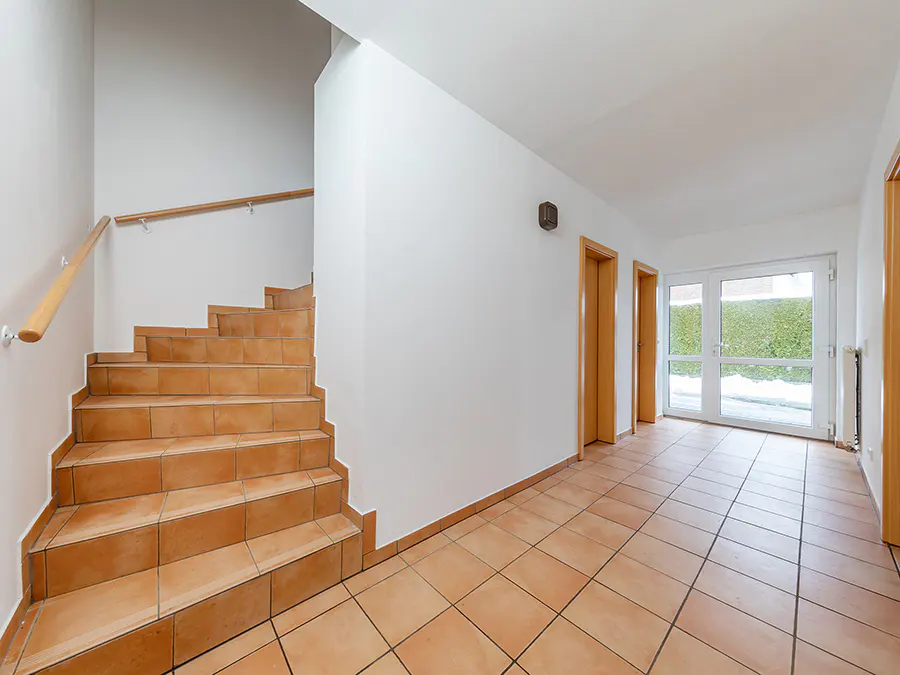 Interior view of a hallway with terracotta tile flooring, a staircase with a wooden handrail, and a glass door leading to the outside.