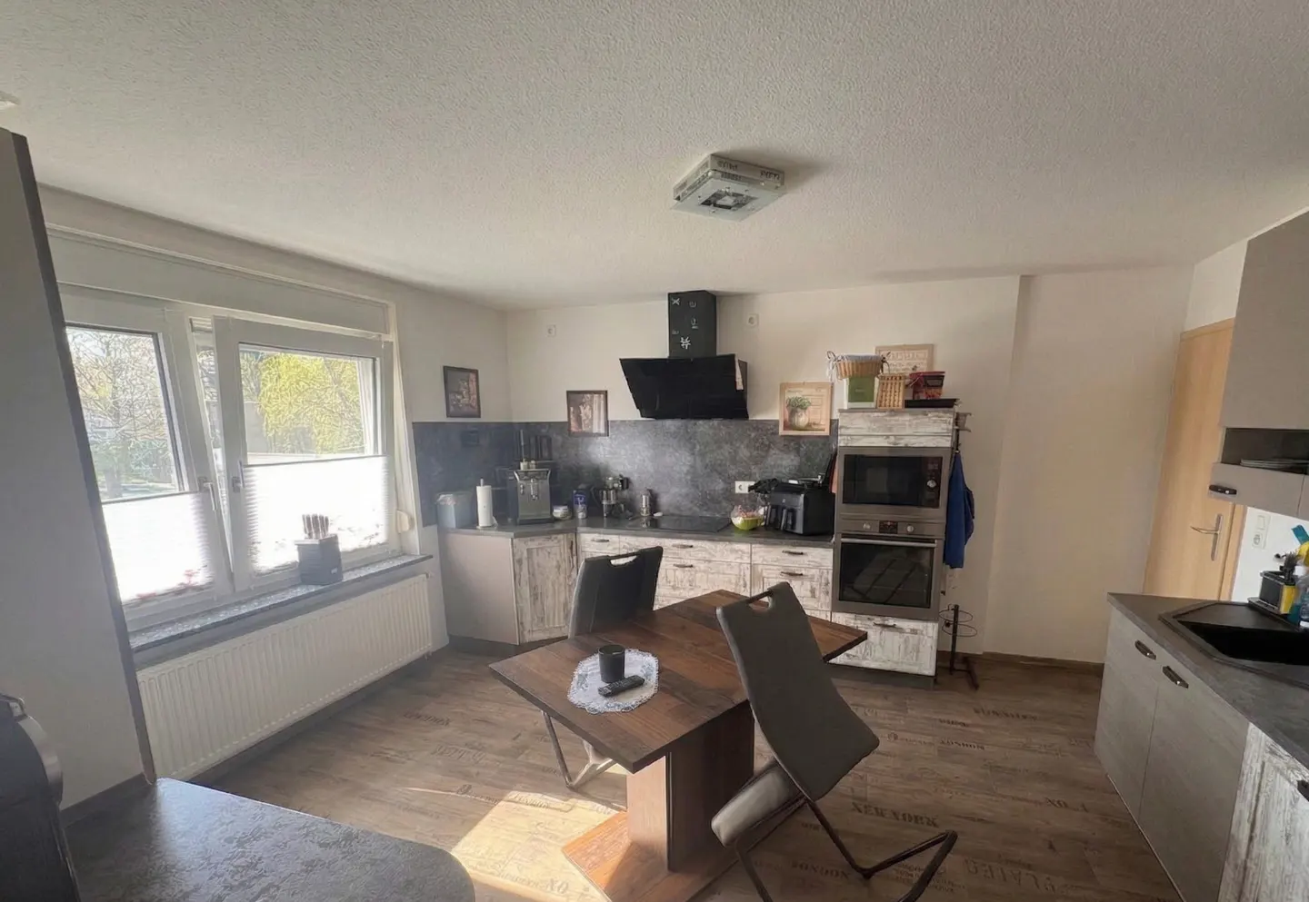 A bright kitchen with a wooden table and chairs in the foreground. The kitchen features white cabinets, a black range hood, and a window with blinds.