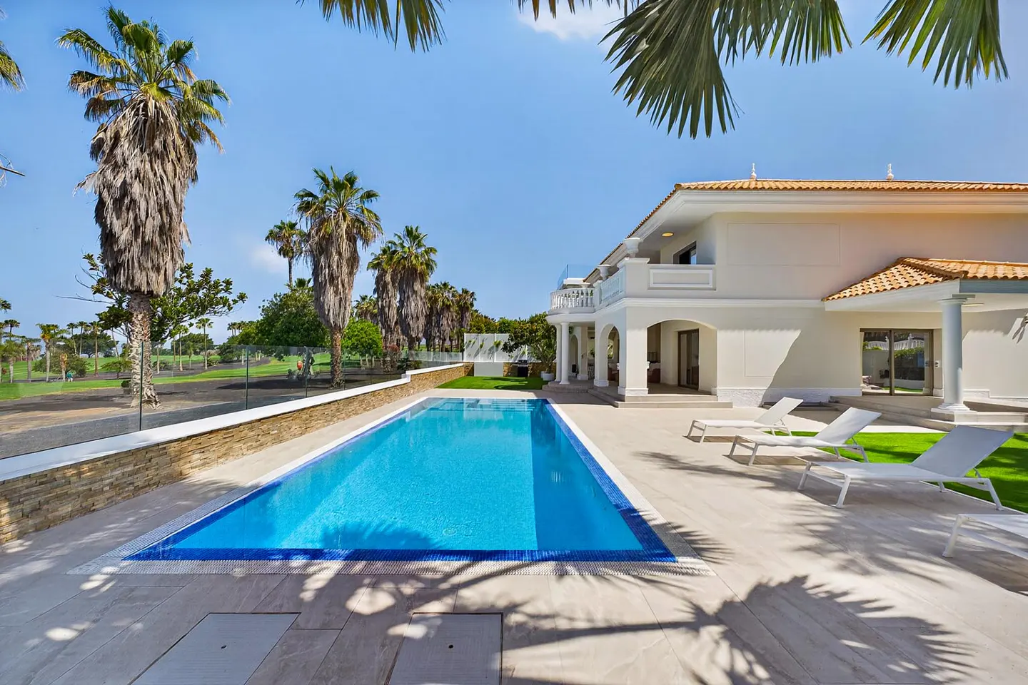 Luxury villa with a blue swimming pool, white lounge chairs, and palm trees against a clear blue sky.
