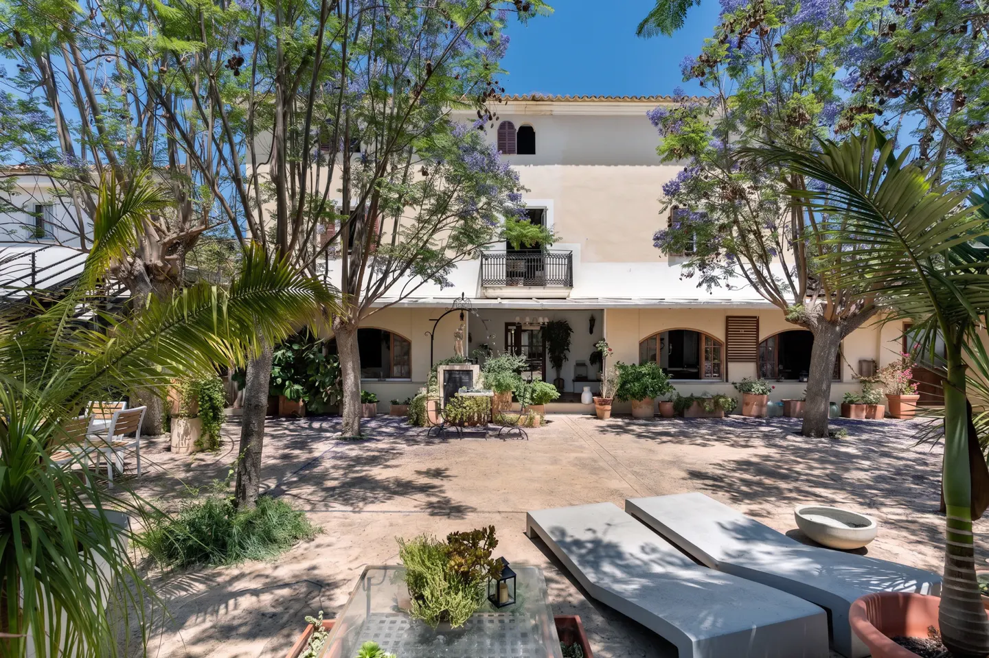 Sunny courtyard of a beige villa with lush trees, potted plants, and two modern lounge chairs on a stone patio.