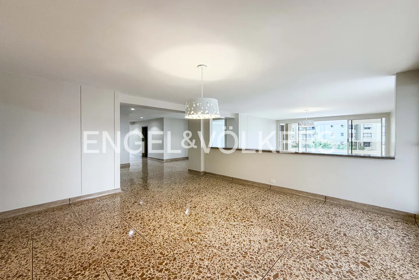 A bright, empty room with terrazzo floors and white walls. A modern chandelier hangs from the ceiling. A large window shows a city view.