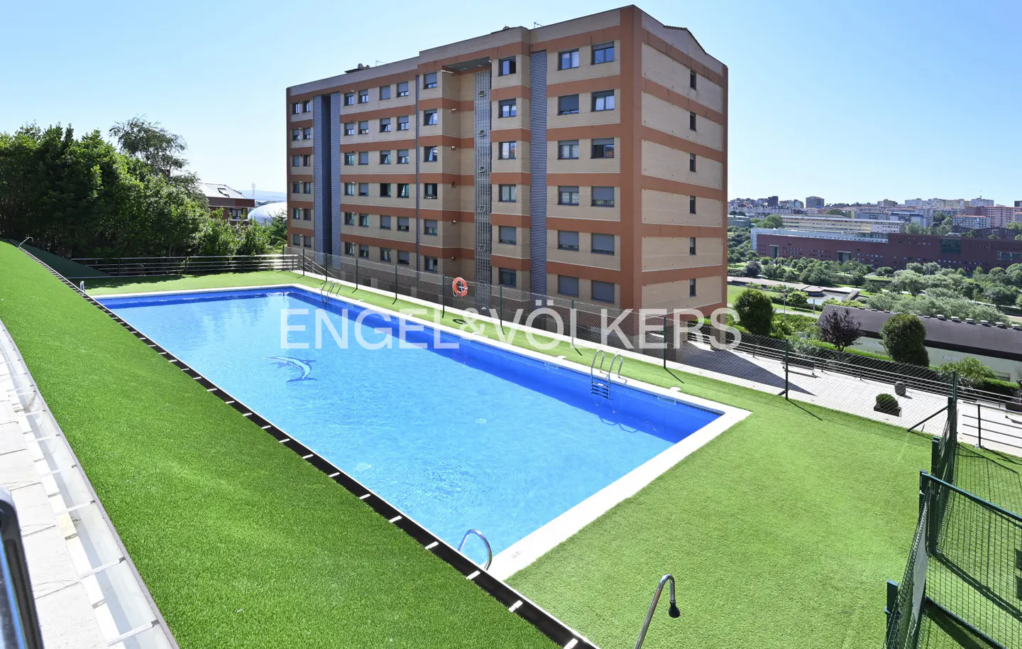 Outdoor pool with blue water and green turf, next to a tall apartment building with beige and brown stripes, under a clear blue sky.