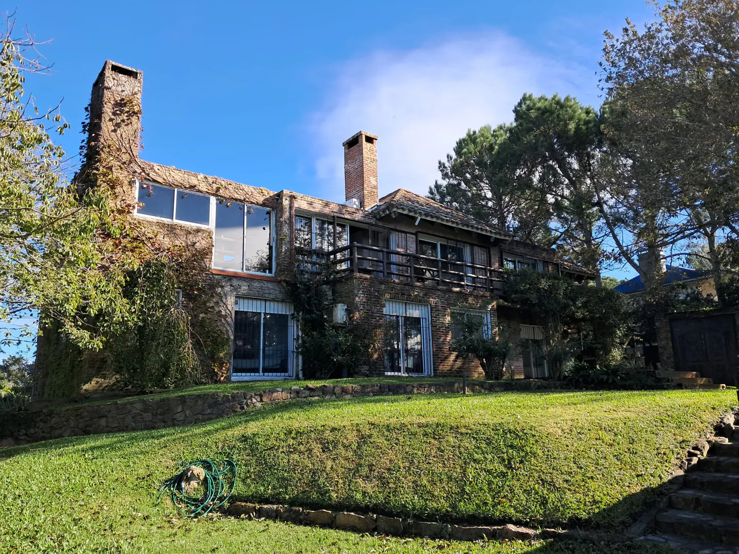Two-story stone house with a chimney, large windows, and a balcony, set on a green lawn under a blue sky.